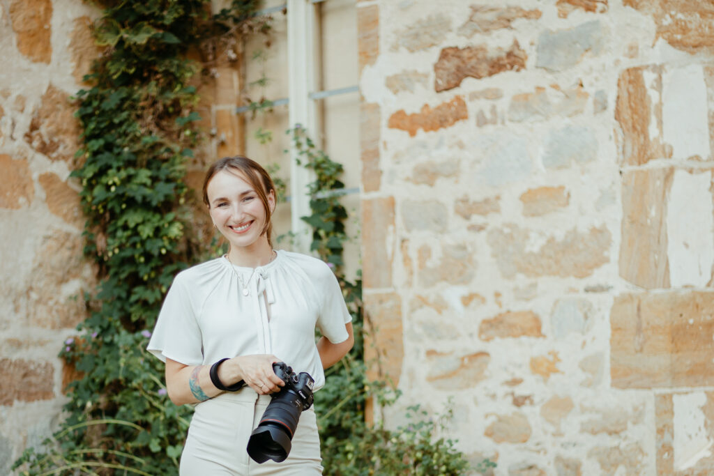 Fotograf Hochzeiten Elopements Portrait Forografin steht mit Kamera in der Handy vor einem mit Efeu bewachsenen Gemäuer