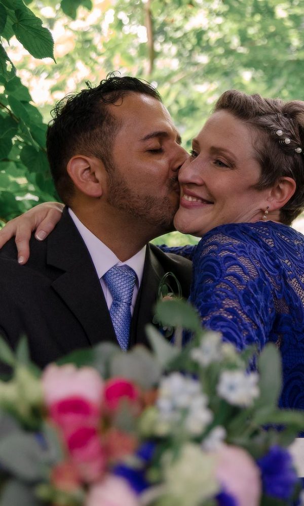 Hochzeitfoto Mit Blumenstraß im Vordergrund Bräutigam küsst Braut auf die Wange
