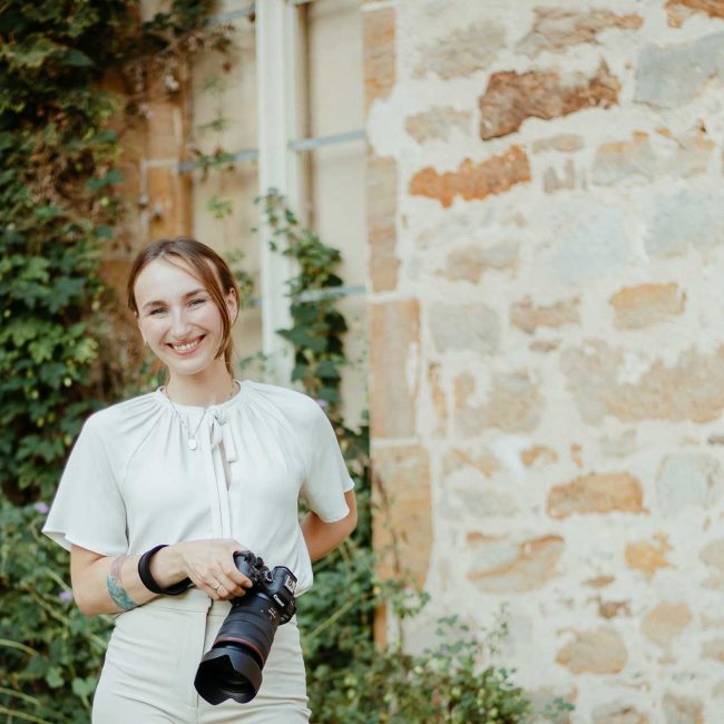 Fotograf Hochzeiten Elopements Portrait Forografin steht mit Kamera in der Handy vor einem mit Efeu bewachsenen Gemäuer