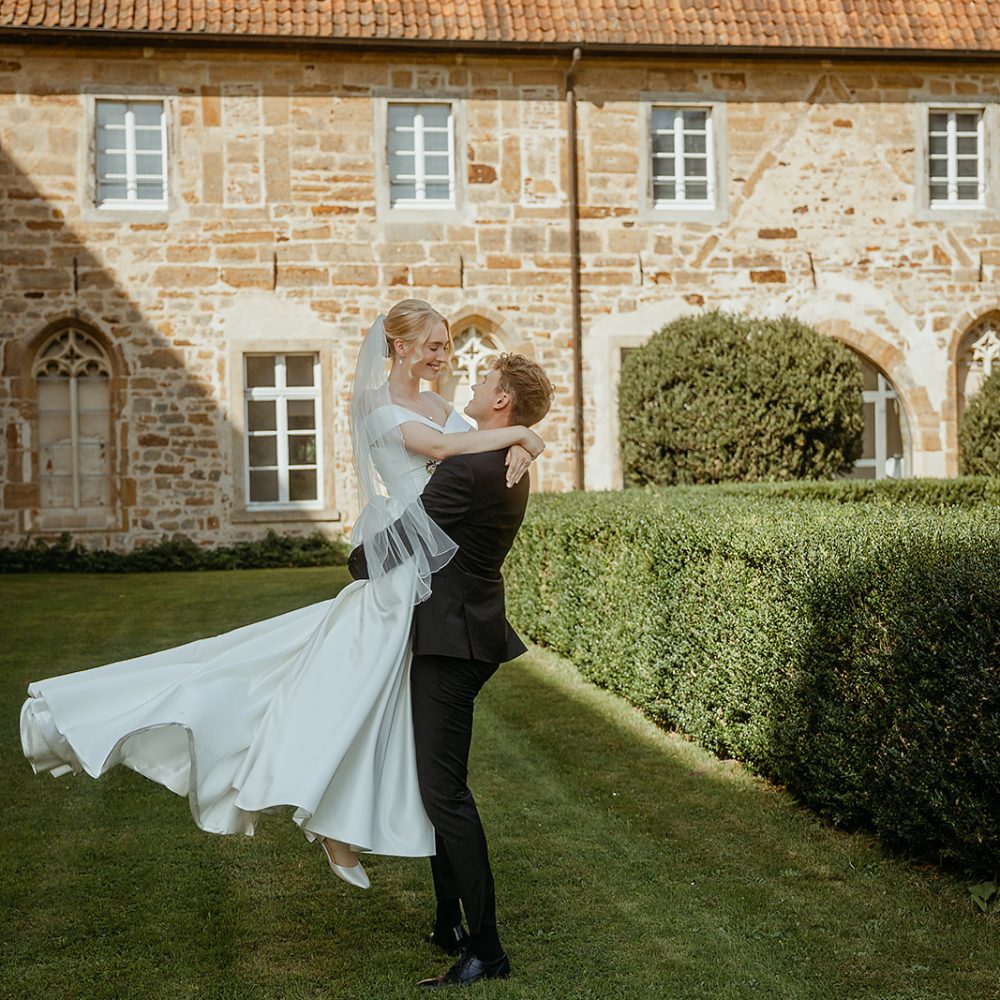 intime exklusive Hochzeitsfotografie im Kloster Bentlage Münster Bräutigam hebt seine Braut mit dem Kloster im Hintergrund